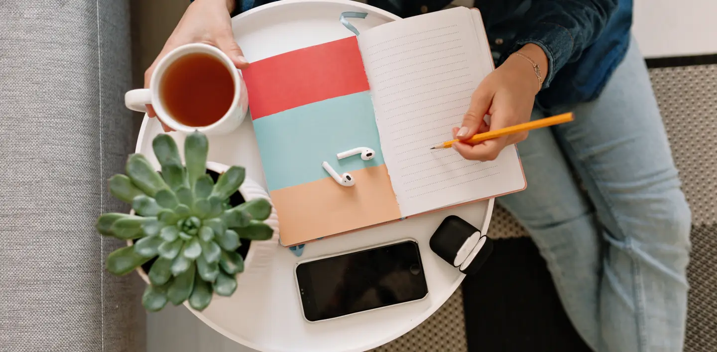 flat-lay-working-table-with-young-womans-hand-writing-notebook-with-smartphone-cup-tea-green-plant