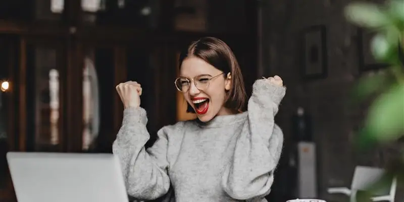 heureuse-femme-a-lunettes-fait-un-geste-gagnant-et-se-rejouit-sincerement-dame-avec-rouge-a-levres-vetue-d-un-pull-gris-regardant-un-ordinateur-portable